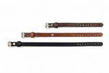 Leather straps in dark brown, medium brown, and black, each with silver-toned metal buckles, laid out vertically on a light textured background.